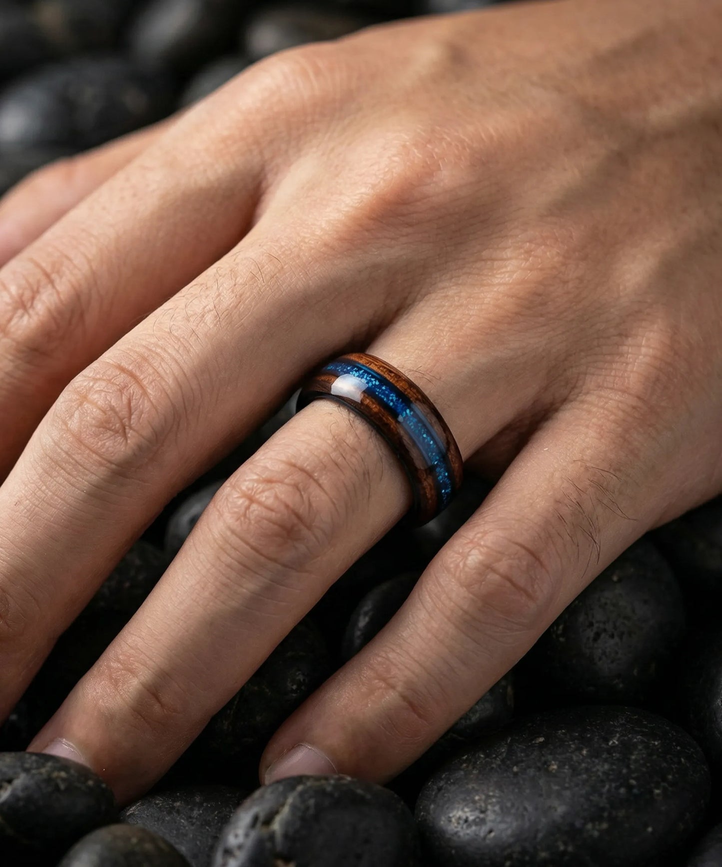 Bague en bois Ark Benetto avec incrustation bleue portée sur une main posée sur des galets noirs, alliance en bois et tungstène au style naturel et élégant.