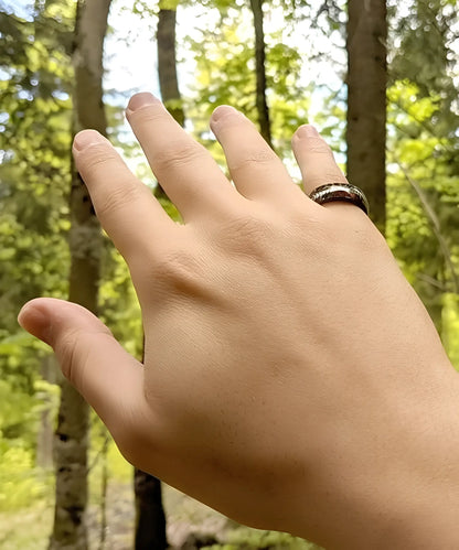 Bague Flèche de Cupidon noire Caprice portée à la main en forêt, anneau en bois et tungstène noir avec flèche argentée, bijou élégant et moderne.