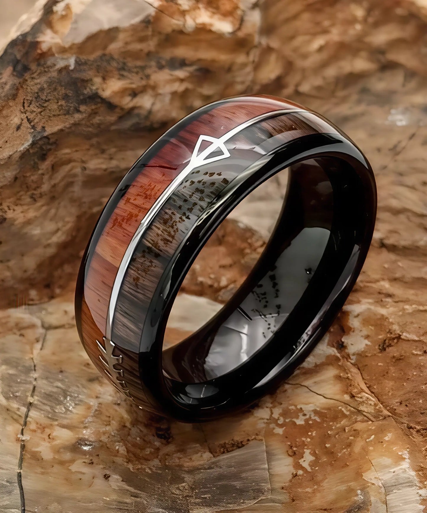 Bague Flèche de Cupidon noire Passion en tungstène noir et bois de koa, gravée d’une flèche argentée, posée sur une surface rocheuse aux tons bruns naturels.