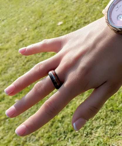 Bague Flèche de Cupidon noire Passion portée à la main d’une femme, en tungstène noir et bois de koa avec flèche argentée, photo en extérieur au soleil.