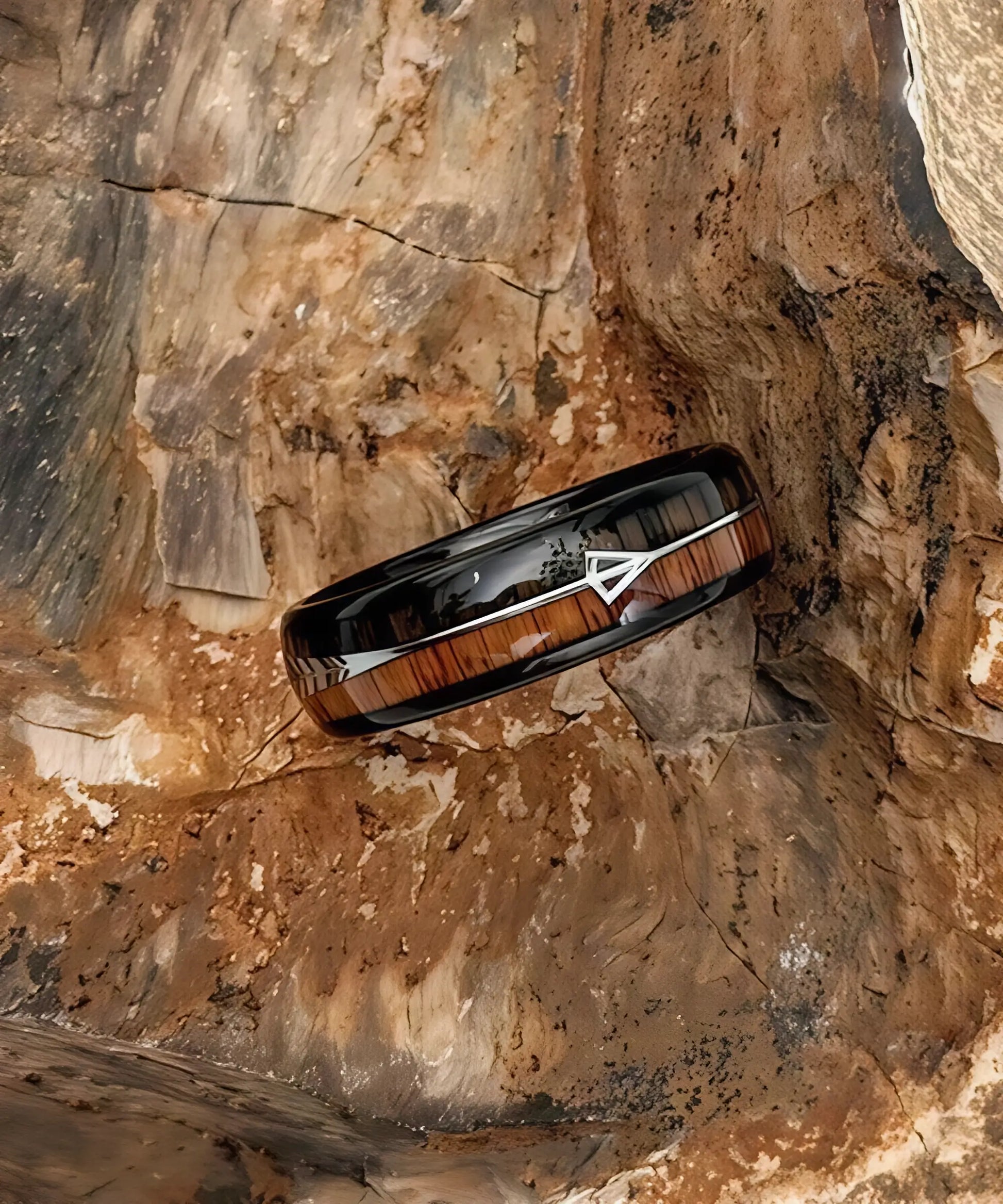 Bague en bois Flèche de Cupidon noire Passion Sim 6 mm, anneau en tungstène noir avec incrustation de bois de koa clair et sombre et motif flèche argentée, posée sur une roche.