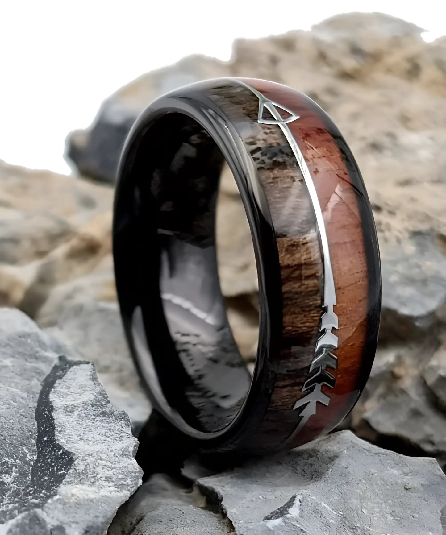 Bague Flèche de Cupidon noire Passion en bois de koa et tungstène noir, posée sur des roches, avec gravure argentée en forme de flèche symbolisant l’amour passionné.