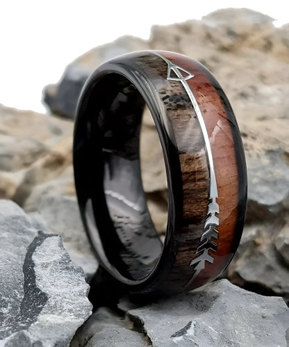 Bague Flèche de Cupidon noire Passion en bois de koa et tungstène noir, posée sur des roches, avec gravure argentée en forme de flèche symbolisant l’amour passionné.