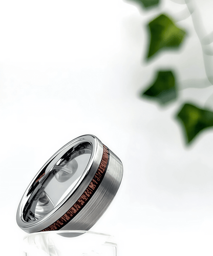 Bague en bois Ultron Ram argent en tungstène finition brossée avec incrustation de bois de koa, présentée sur support studio avec feuillage décoratif